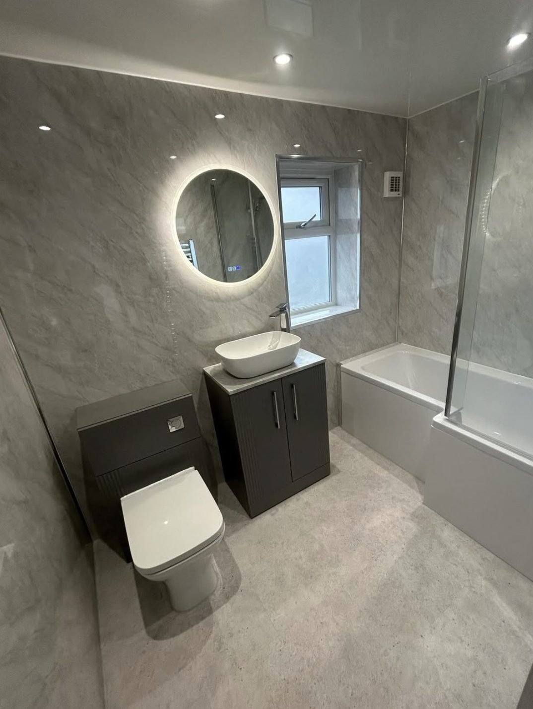 Grey marble-effect panelled bathroom with backlit round mirror, vanity unit and bath