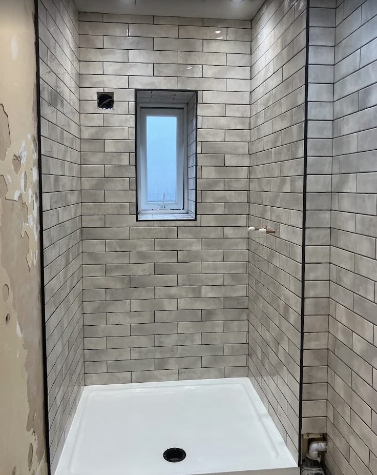 Shower enclosure part way through tiling with concrete effect metro tiles around a window reveal
