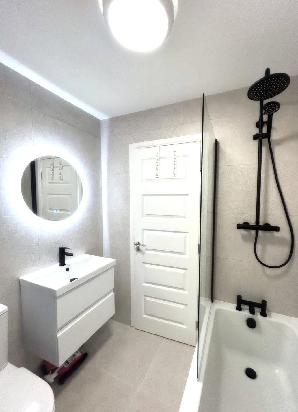 White bathroom with large format tiles, matt black shower over bath and backlit round mirror